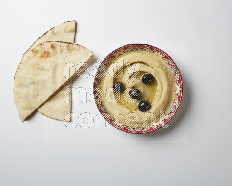 Hummus in a red plate with patterns garnished with black olives on a white background
