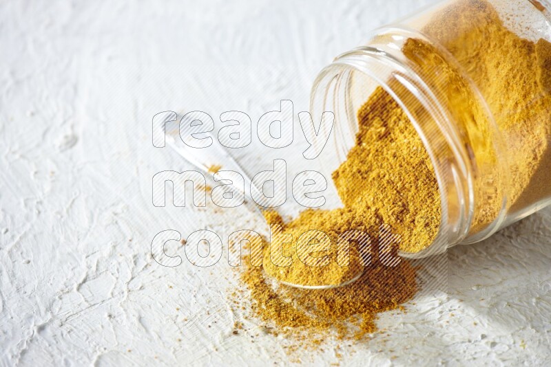 A flipped glass spice jar and a metal spoon full of turmeric powder and powder spilled out of it on textured white flooring