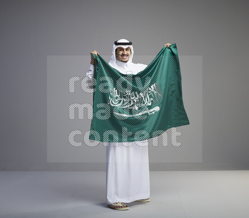 رجل سعودي يرتدي ثوب ابيض وشماغ يحمل العلم السعودي
