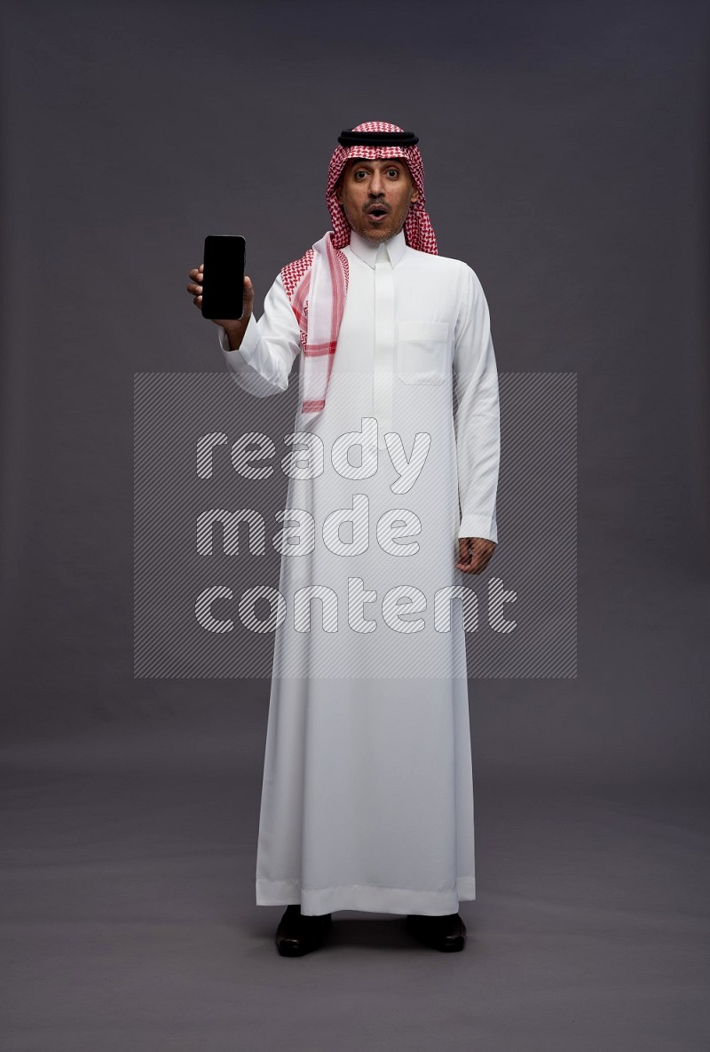 Saudi man wearing thob and shomag standing showing phone to camera on gray background
