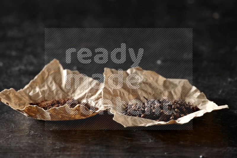 Cloves powder and cloves in 2 crumpled pieces of paper on a textured black flooring