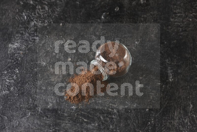 A flipped glass spice jar full of cloves powder and powder came out of it on textured black flooring