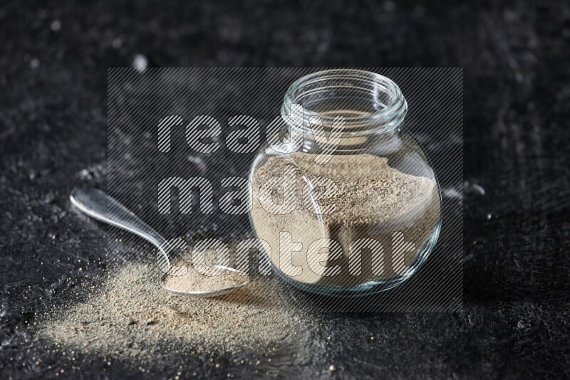 Herbal glass jar and metal spoon full of white pepper powder on textured black flooring