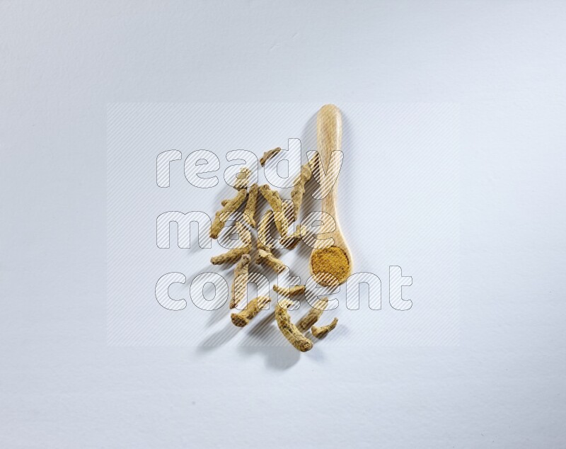 ملعقة خشبية ممتلئة ببودرة الكركم مع أصابع الكركم المجففة بجانبها على خلفية بيضاء