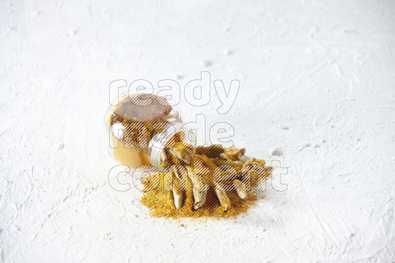 وعاء زجاجي للتوابل مقلوب ممتلئ ببودرة الكركم والبودرة تناثرت منه مع أصابع الكركم المجففة على خلفية بيضاء