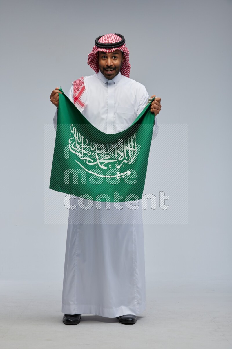 Saudi man Wearing Thob and shomag standing holding Saudi flag on Gray background