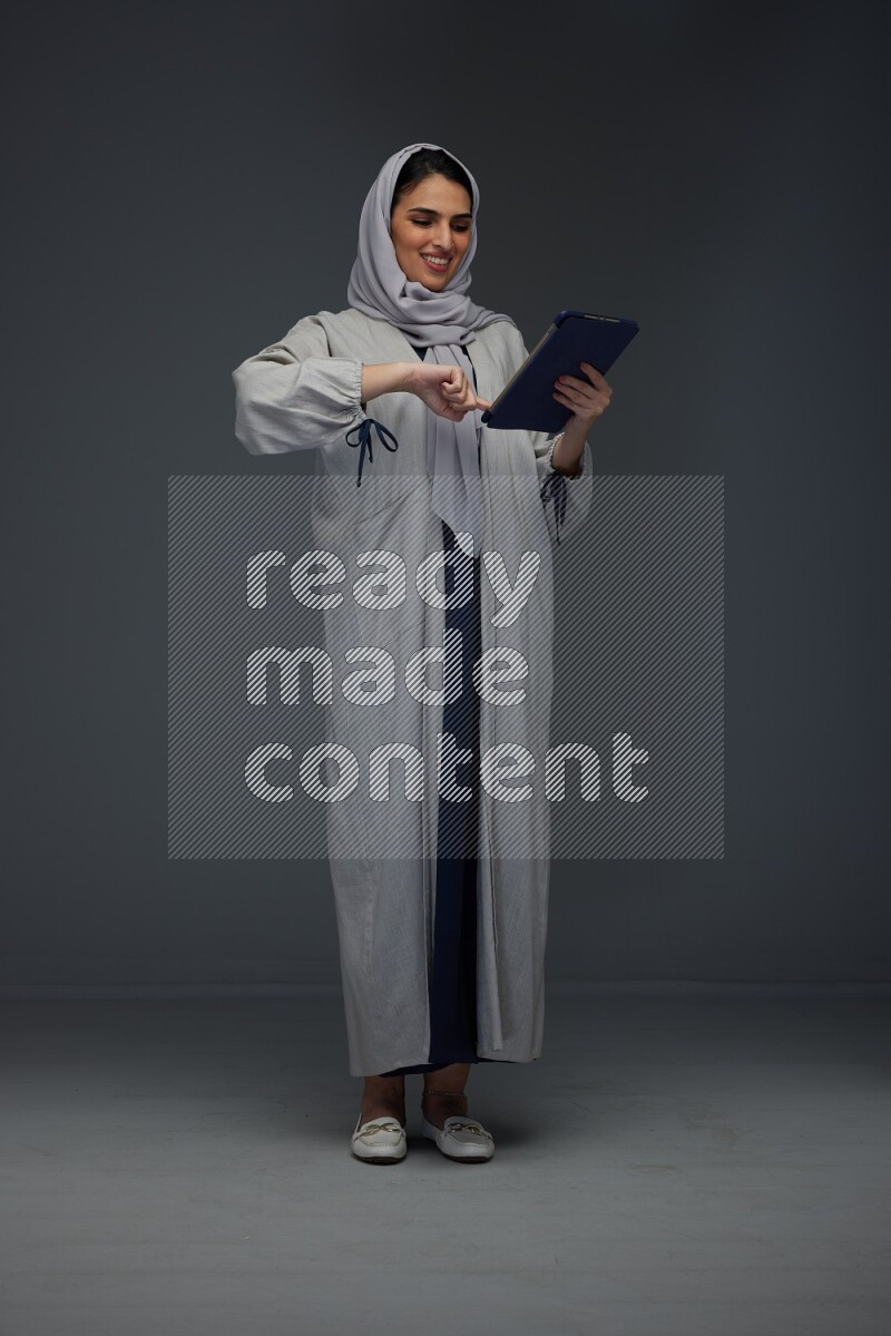 A Saudi woman wearing a light gray Abaya and head scarf standing holding a phone and pointing with the other hand eye level on a grey background