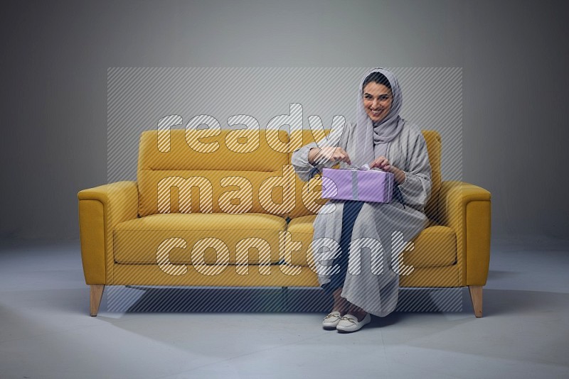 A Saudi woman wearing a light gray Abaya and head scarf sitting on a yellow sofa holding a gift box beside multi color gift boxes eye level on a grey background