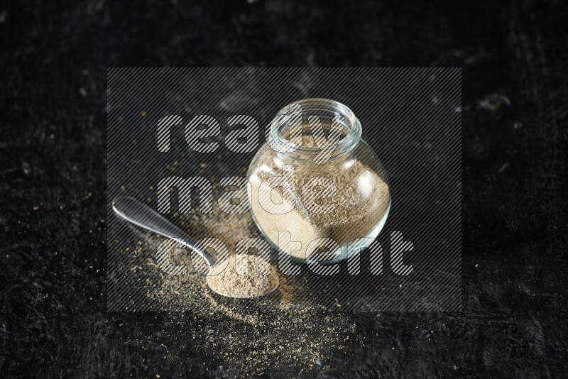 A glass spice jar and metal spoon full of cardamom powder on textured black flooring