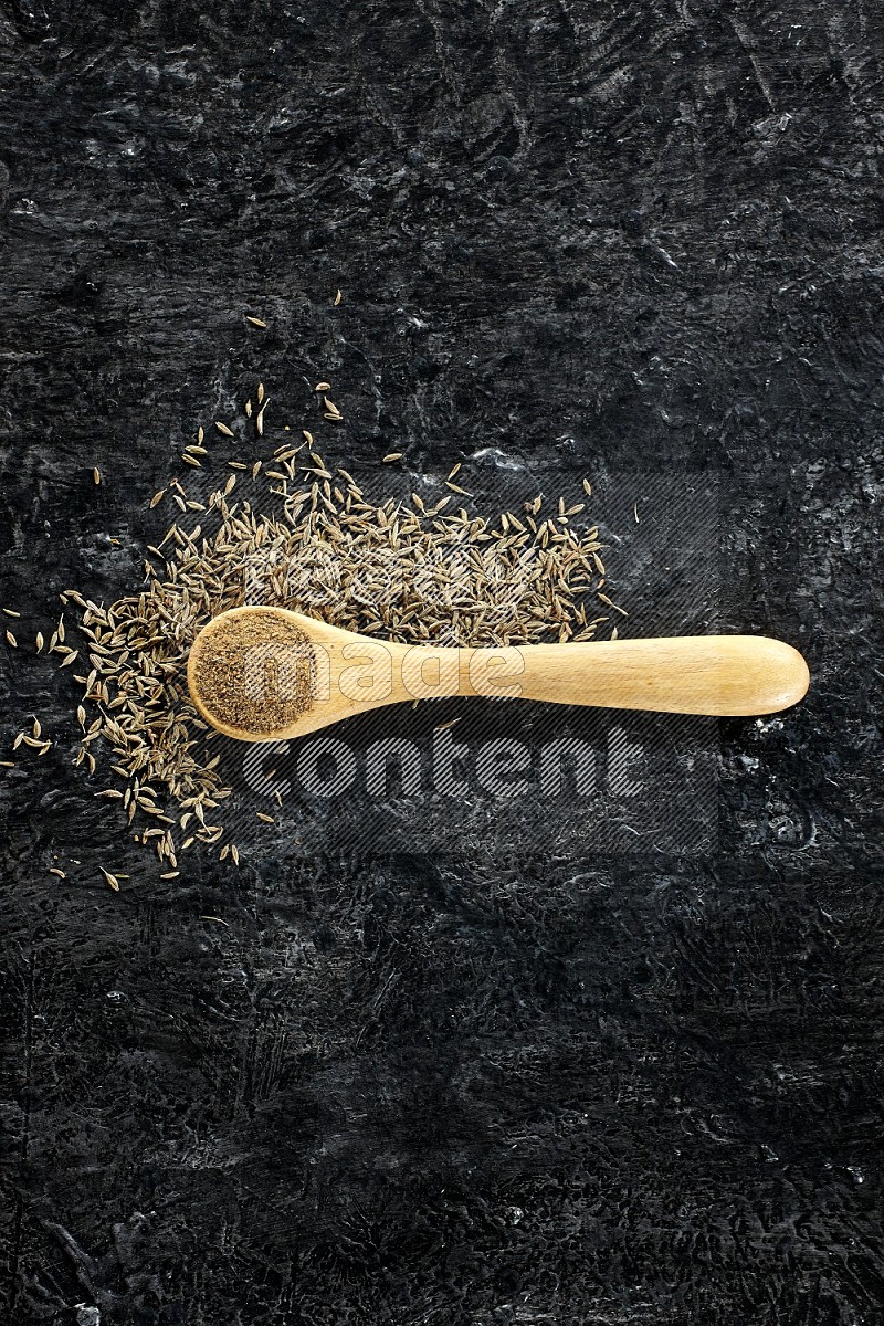A wooden spoon full of cumin powder and cumin seeds spreaded on textured black flooring