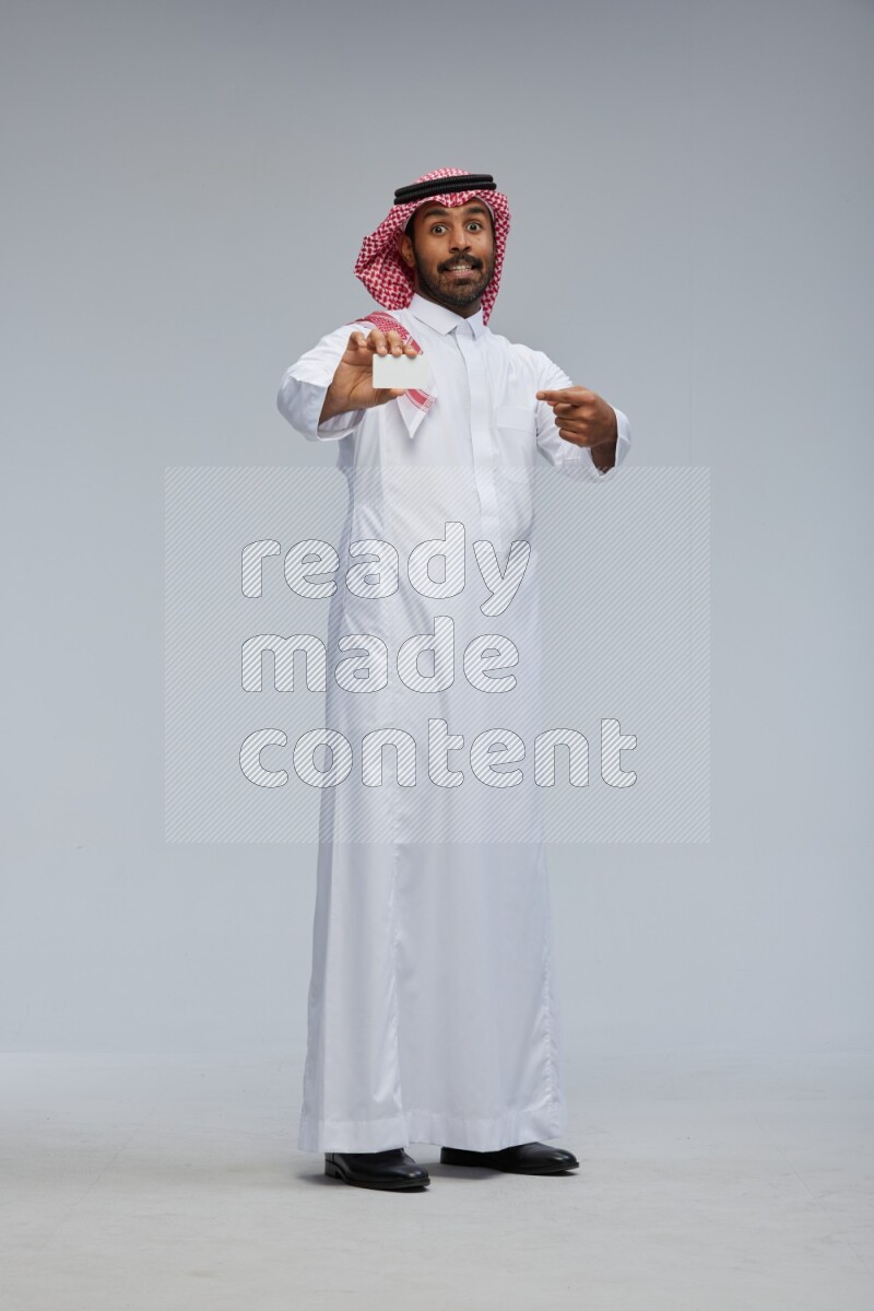 Saudi man wearing thob and shomag standing holding ATM card on gray background