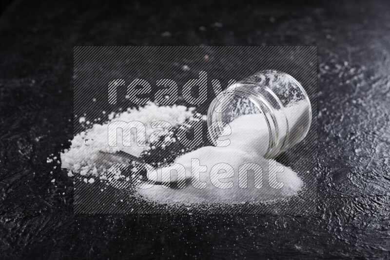 A glass jar full of table salt with some sea salt crystals beside it on a black background