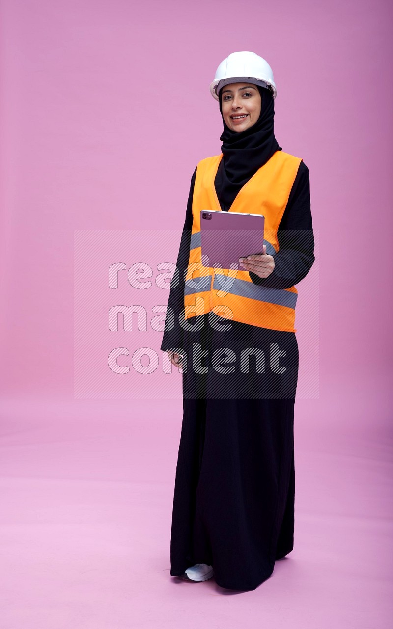 Saudi woman wearing Abaya with engineer vest and helmet standing working on tablet on pink background