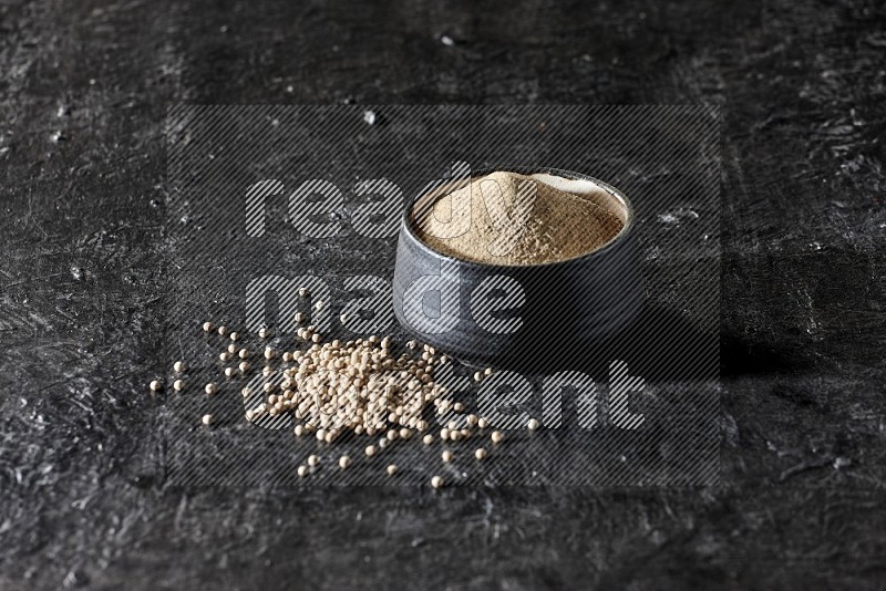 Black pottery bowl full of white pepper powder with white pepper beads on textured black flooring