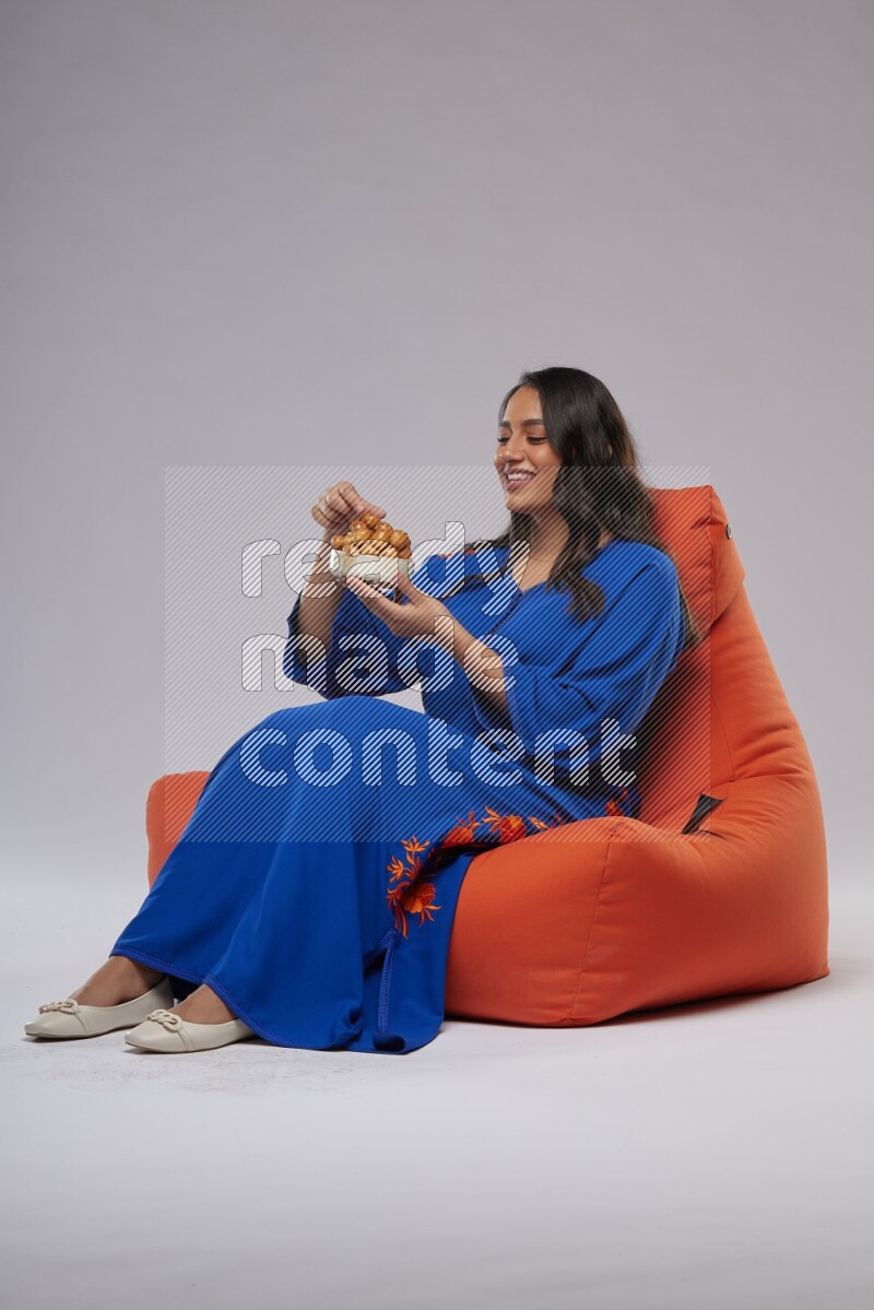 A Woman sitting on an orange beanbag wearing Jalabeya holding a plate of luqaimat