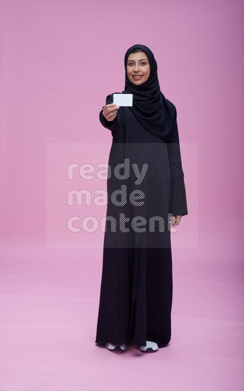 Saudi woman wearing Abaya standing holding ATM card on pink background