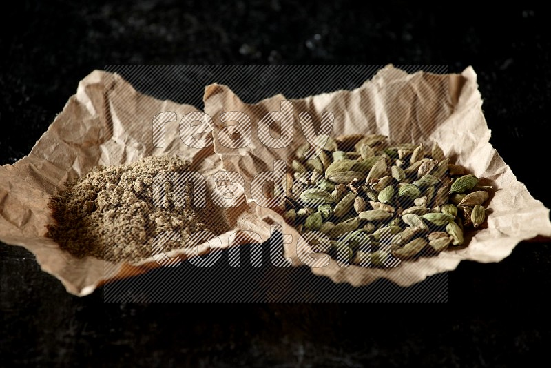 Cardamom seeds and cardamom powder in 2 crumpled pieces of paper on textured black flooring