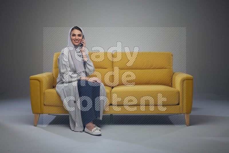 A Saudi woman wearing a light gray Abaya and head scarf sitting on a yellow sofa and talking in the phone eye level on a grey background
