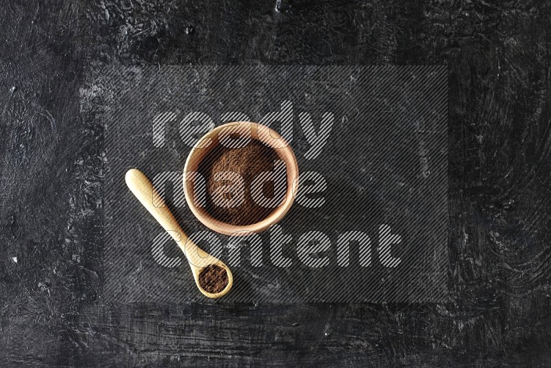 A wooden bowl and wooden spoon full of cloves powder on textured black flooring