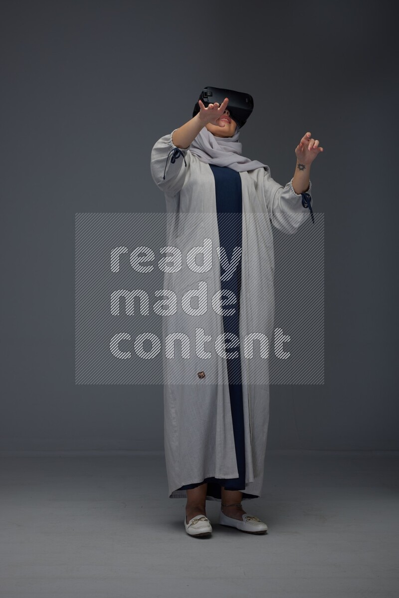 A Saudi woman wearing a light gray Abaya and head scarf standing and wearing VR eye level on a grey background