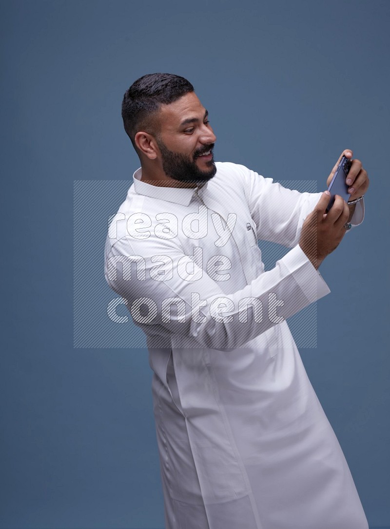 A man Playing a Game on his Smartphone  on Blue Background wearing Saudi Thob