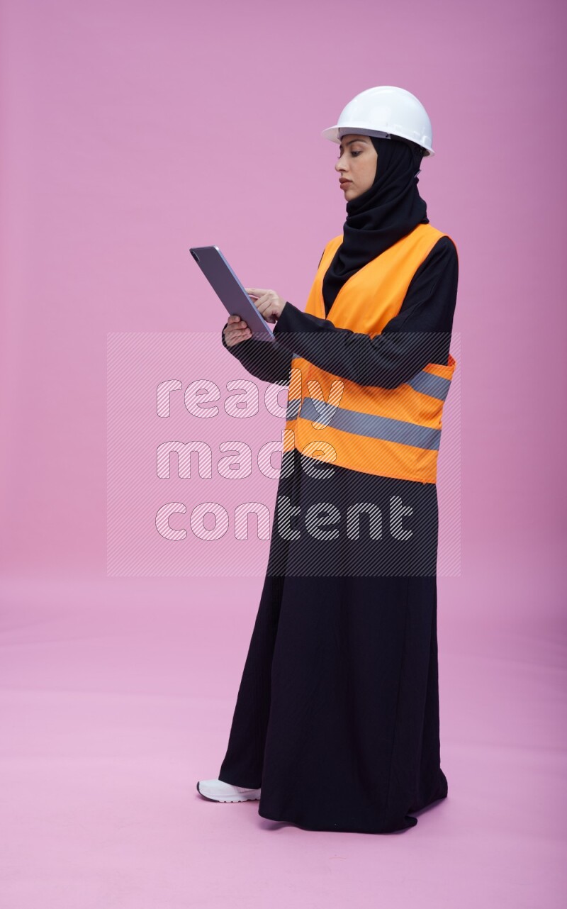 Saudi woman wearing Abaya with engineer vest and helmet standing working on tablet on pink background