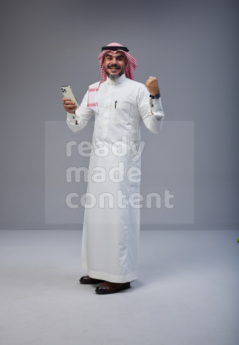 Saudi man Wearing Thob and red Shomag standing texting on phone on Gray background