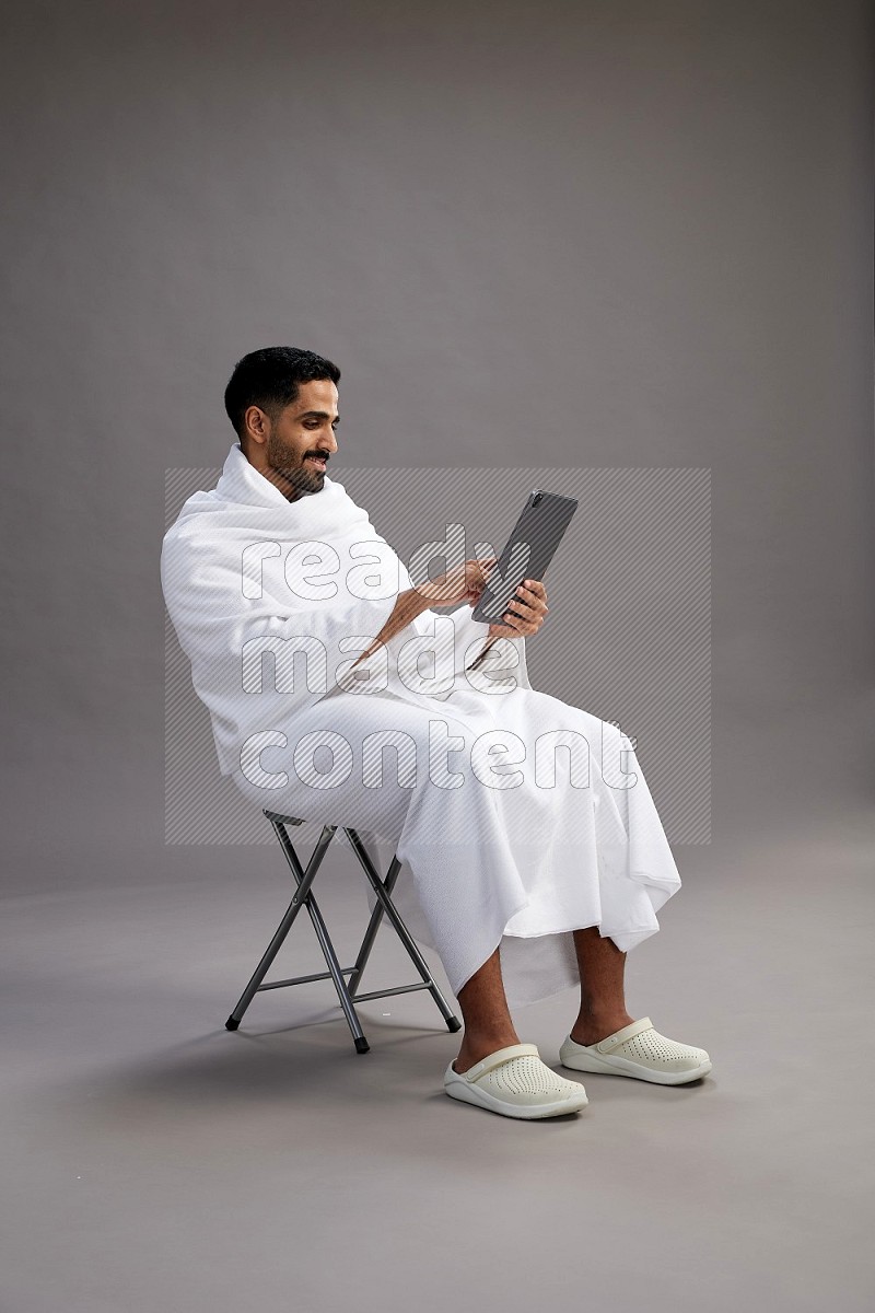 A man wearing Ehram sitting on chair holding a tablet on gray background