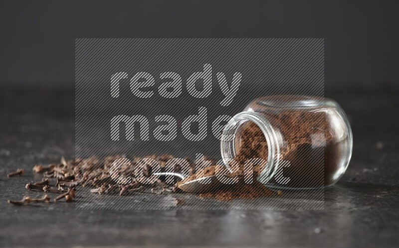 A flipped glass spice jar and a metal spoon full of cloves powder with cloves spread on textured black flooring