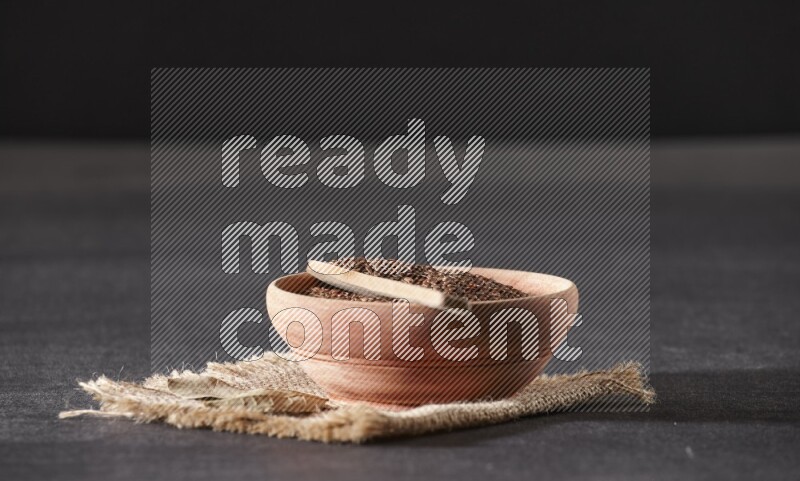 A wooden bowl full of flaxseeds with wooden spoon full of the seeds on it on burlap fabric on a black flooring