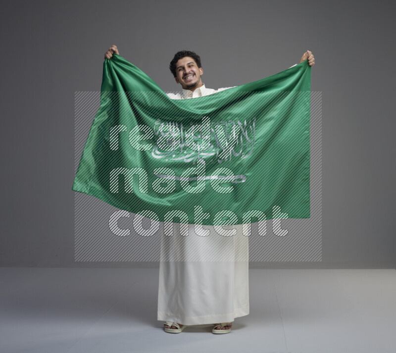 A Saudi man standing wearing thob holding big Saudi flag on gray background