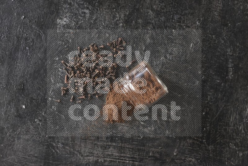 وعاء زجاجي مقلوب ممتلئ ببودرة القرنفل مع حبوب القرنفل المتناثرة على أرضية سوداء