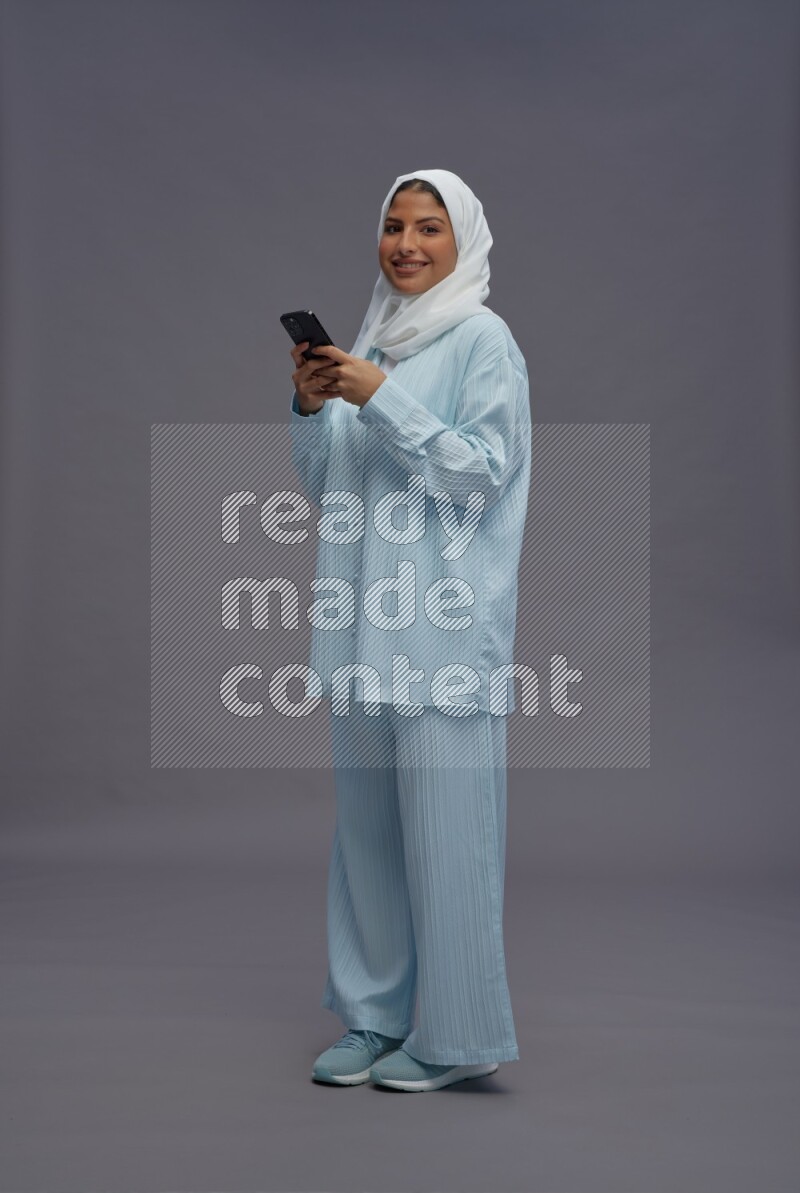 Saudi woman wearing hijab clothes standing texting on phone on gray background