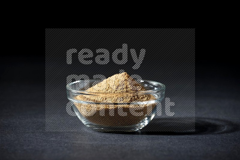 A glass bowl full of cumin powder on black flooring
