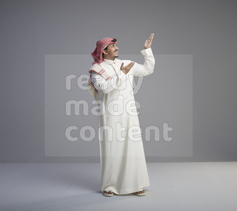 A Saudi man standing wearing thob and red shomag interacting with the camera on gray background