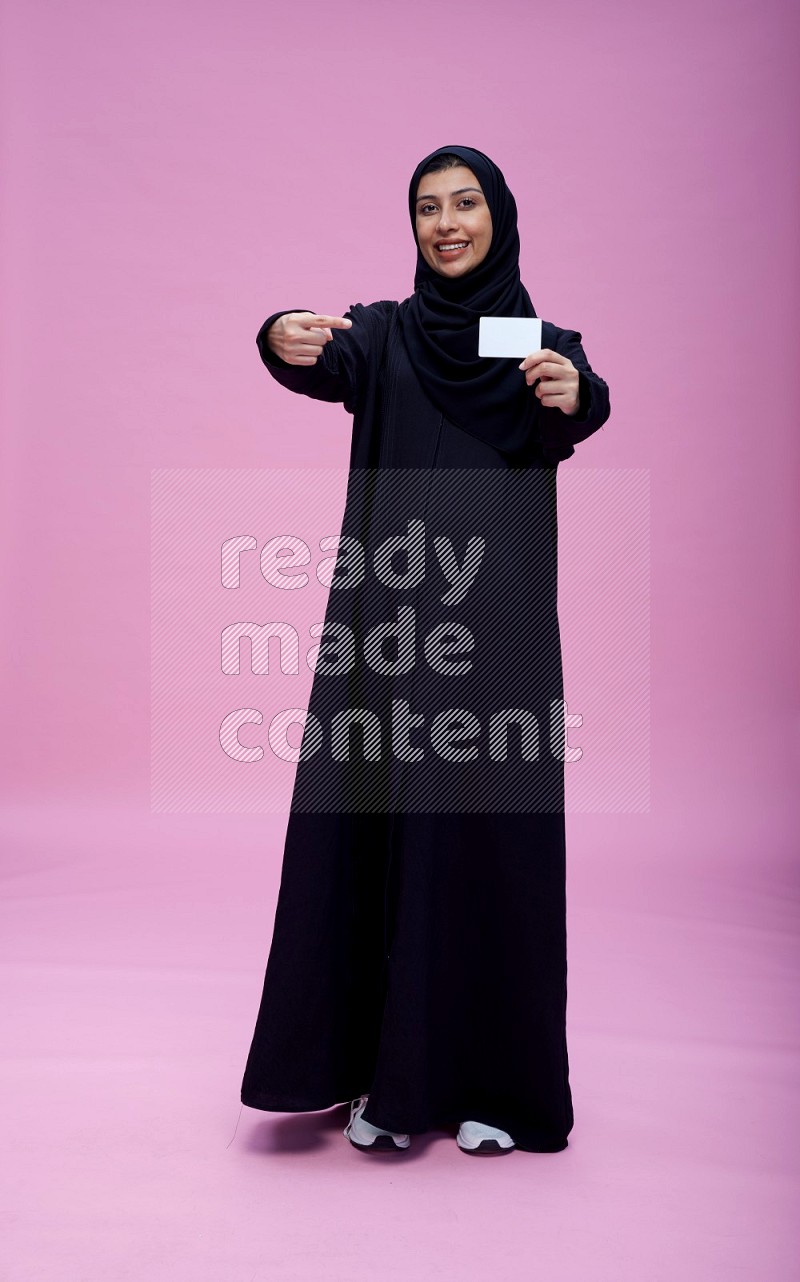 Saudi woman wearing Abaya standing holding ATM card on pink background