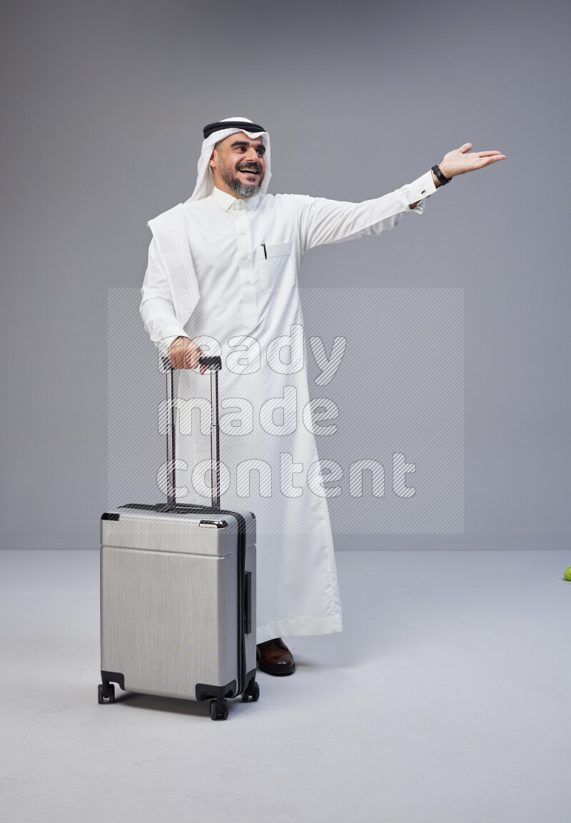 Saudi man wearing Thob and white Shomag standing holding Travel bag on Gray background