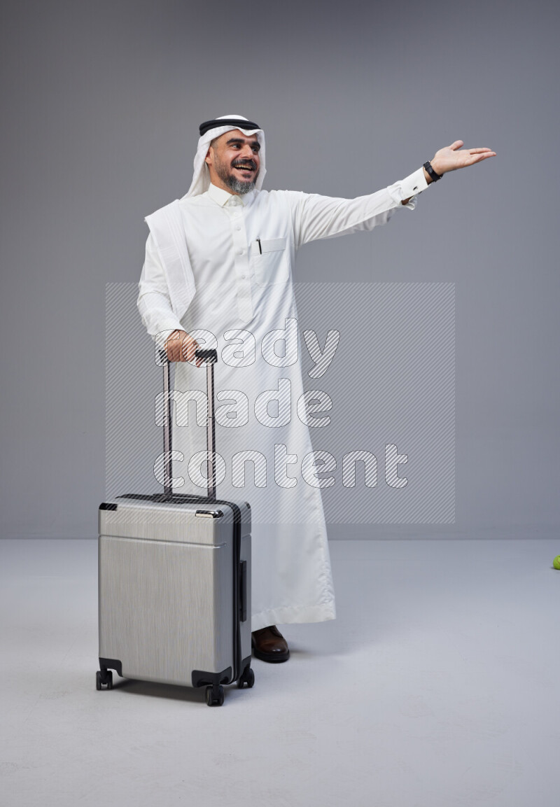Saudi man wearing Thob and white Shomag standing holding Travel bag on Gray background