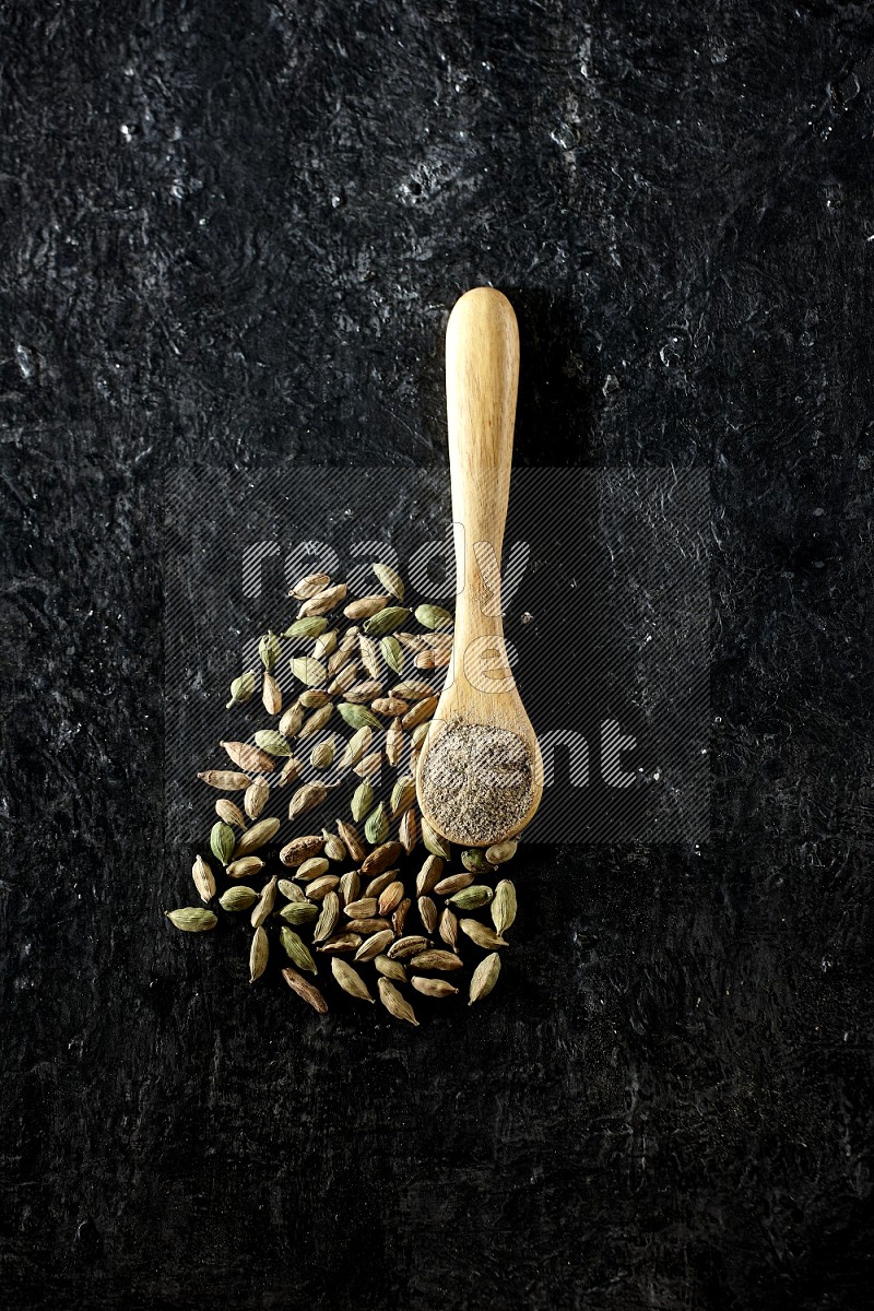 A wooden spoon full of cardamom powder and cardamom seeds next of it on textured black flooring