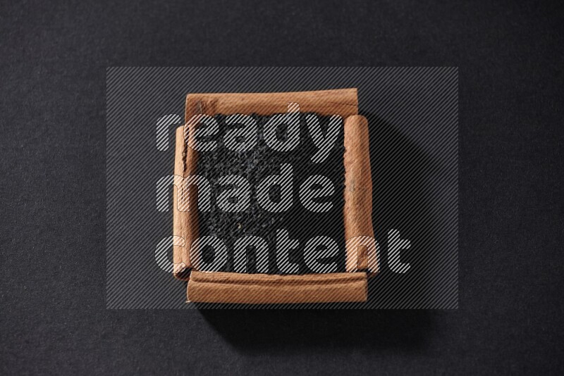 أربعة من أعواد القرفة مكونة شكل مربع بداخلها حبة البركة علي خلفية سوداء