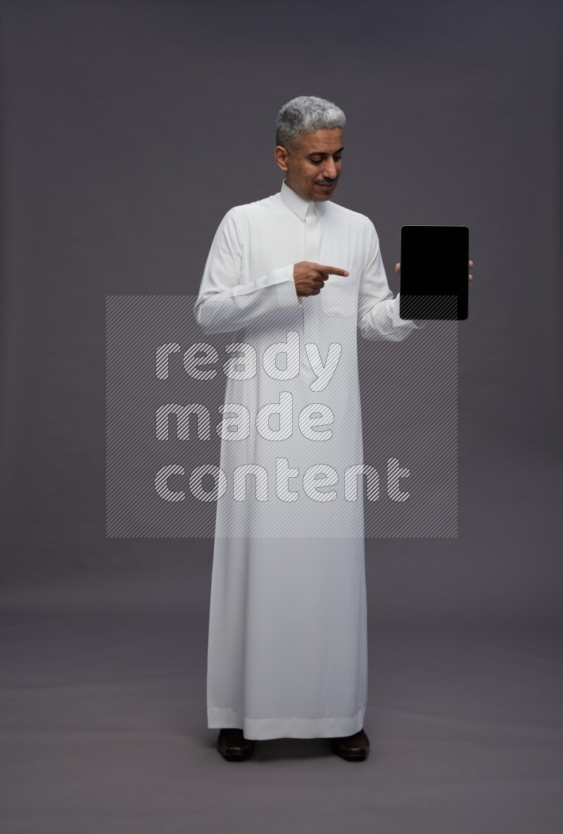 Saudi man wearing thob standing showing tablet to camera on gray background