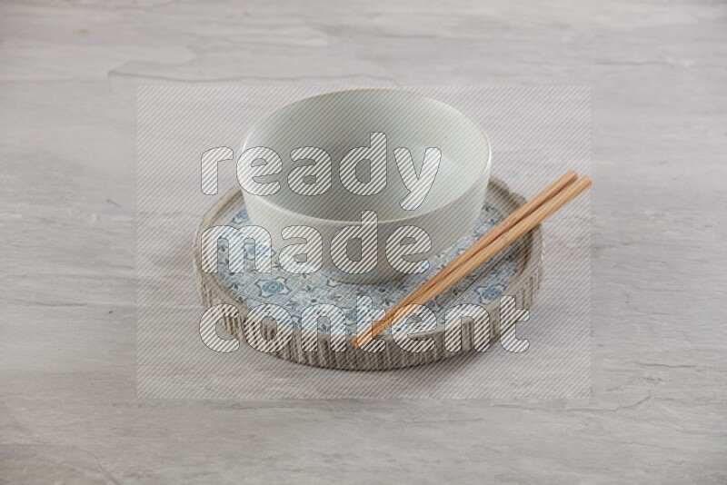 white pottery round bowl on top of multi color round ceramic plate and chopsticks, on grey textured countertop