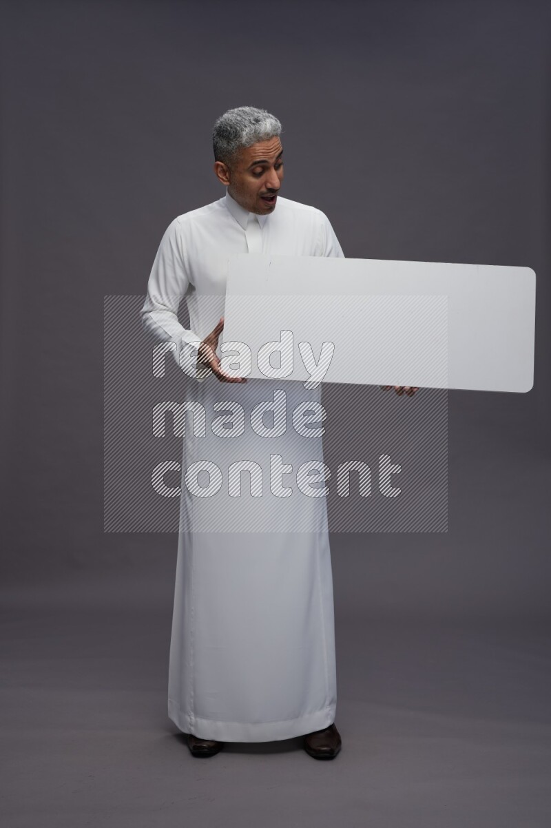Saudi man wearing thob standing holding board on gray background