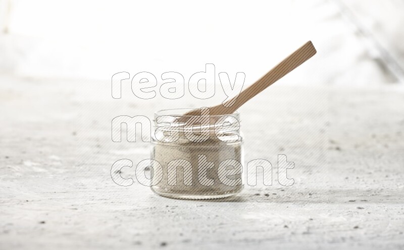A glass jar and wooden spoon full of white pepper powder on textured white flooring