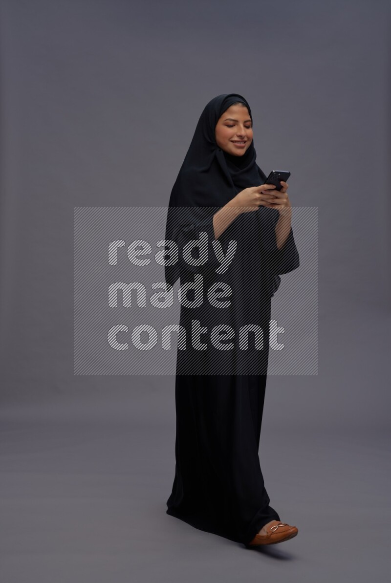 Saudi woman wearing Abaya standing texting on phone on gray background