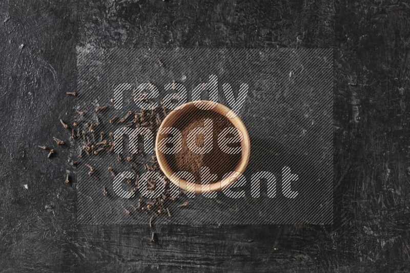 A wooden bowl full of cloves powder on a textured black flooring