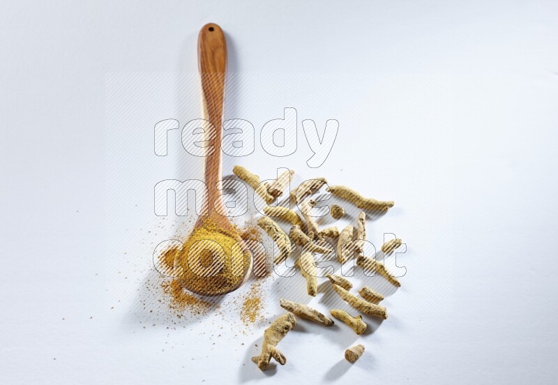 A wooden ladle full of turmeric powder and dried turmeric fingers beside it on white flooring
