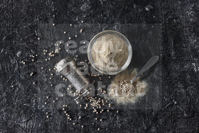 A glass bowl full of white pepper powder with pepper beads, a metal grinder and a metal spoon on textured black flooring