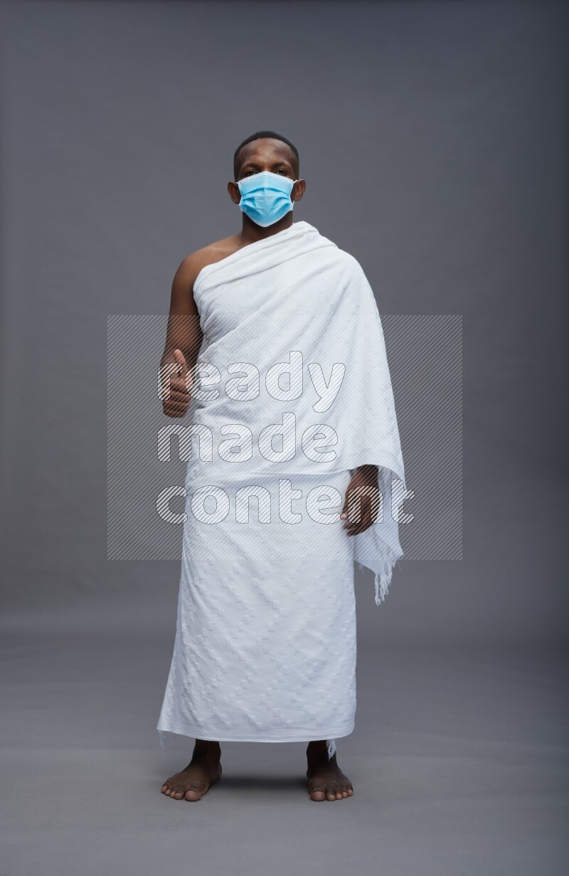 A man wearing Ehram with face mask Standing Interacting with the camera on gray background