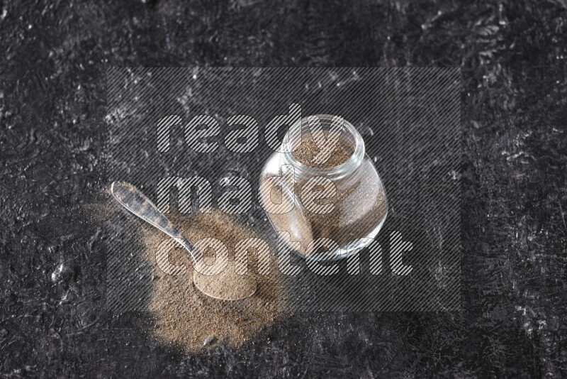 A glass spice jar full of black pepper powder and a metal spoon full of powder on textured black flooring